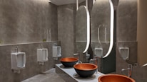 A modern bathroom with a sophisticated design featuring large oval mirrors with built-in lighting above elegant stone countertops. The room includes several urinals along one wall, each separated by partitions, and two round vessel sinks with sleek faucets. The wall tiles and flooring have a textured gray finish, contributing to the contemporary aesthetic.