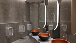 A modern bathroom with a sophisticated design featuring large oval mirrors with built-in lighting above elegant stone countertops. The room includes several urinals along one wall, each separated by partitions, and two round vessel sinks with sleek faucets. The wall tiles and flooring have a textured gray finish, contributing to the contemporary aesthetic.