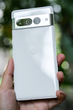 A hand holds a sleek, white smartphone with a glossy finish. The device features a metallic camera strip with multiple lenses and sensors on the back. The background is softly blurred with hints of greenery.