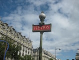 A classic Parisian metro sign with a red background and white lettering is prominently displayed. The sign is attached to a vintage-style green pole topped with a spherical light. In the background, ornate Haussmann-style buildings with mansard roofs and dormer windows are visible under a partly cloudy sky.