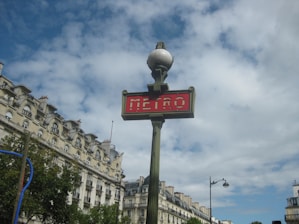 A classic Parisian metro sign with a red background and white lettering is prominently displayed. The sign is attached to a vintage-style green pole topped with a spherical light. In the background, ornate Haussmann-style buildings with mansard roofs and dormer windows are visible under a partly cloudy sky.