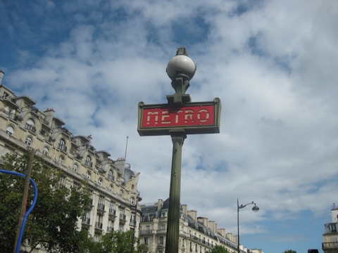 A classic Parisian metro sign with a red background and white lettering is prominently displayed. The sign is attached to a vintage-style green pole topped with a spherical light. In the background, ornate Haussmann-style buildings with mansard roofs and dormer windows are visible under a partly cloudy sky.