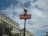A classic Parisian metro sign with a red background and white lettering is prominently displayed. The sign is attached to a vintage-style green pole topped with a spherical light. In the background, ornate Haussmann-style buildings with mansard roofs and dormer windows are visible under a partly cloudy sky.