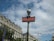 A classic Parisian metro sign with a red background and white lettering is prominently displayed. The sign is attached to a vintage-style green pole topped with a spherical light. In the background, ornate Haussmann-style buildings with mansard roofs and dormer windows are visible under a partly cloudy sky.