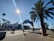 A daytime scene featuring the entrance to a pier with palm trees lining the walkway. The sky is clear and the sun is prominently visible, casting shadows on the pavement. People are walking around, enjoying the sunny weather. The sign prominently displays the words 'Santa Monica Yacht Harbor Sport Fishing Boating Cafes'.