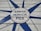 A painted sign on wooden planks featuring a starburst design with the words 'Santa Monica Pier' prominently displayed in the center. The text is surrounded by a blue circle and radiating blue lines extending outwards. Additional text includes 'another day in paradise' and 'est. 2018 by Mimi Gaudet'.