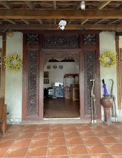 An intricately carved wooden doorway leads into a rustic interior with exposed wooden beams. The door frame is decorated with elaborate floral carvings and two wooden giraffe sculptures stand to the right. Yellow wreaths adorn the doorway on both sides. Inside, there is a beige tiled floor and various furniture pieces including a desk and chairs, with some framed artwork on the wall.