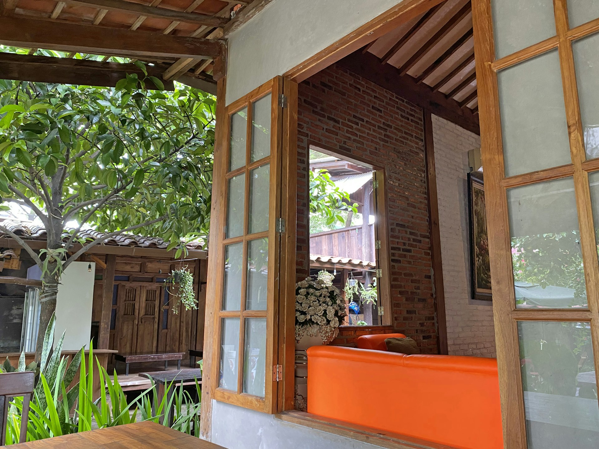 A sunlit living room featuring warm orange-red accents, natural wood textures, and large windows opening to a lush garden, embodying studio bodhi’s harmony of nature and design.