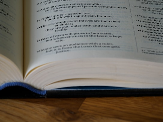 A close-up of an open book, with text printed on cream-colored pages. The book is laid on a wooden surface and partially bound with a dark blue cover, showing clear lines of text.