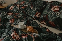 A cozy bed features floral-patterned dark bedding, with a wooden tray resting on it holding a white mug and a croissant. A book is visible on a bedside table in the background.