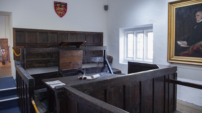 A historic courtroom setting featuring a wooden witness stand or dock with a surrounding fence and podium. The room is adorned with wood paneling on the walls and a framed portrait hanging to the right. A large window allows natural light to illuminate the space. A coat of arms hangs on one of the walls.