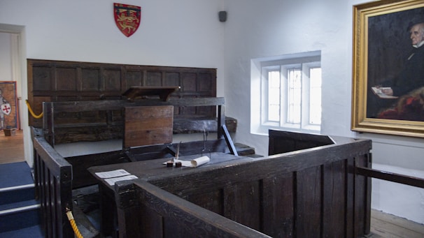 A historic courtroom setting featuring a wooden witness stand or dock with a surrounding fence and podium. The room is adorned with wood paneling on the walls and a framed portrait hanging to the right. A large window allows natural light to illuminate the space. A coat of arms hangs on one of the walls.