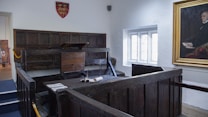 A historic courtroom setting featuring a wooden witness stand or dock with a surrounding fence and podium. The room is adorned with wood paneling on the walls and a framed portrait hanging to the right. A large window allows natural light to illuminate the space. A coat of arms hangs on one of the walls.