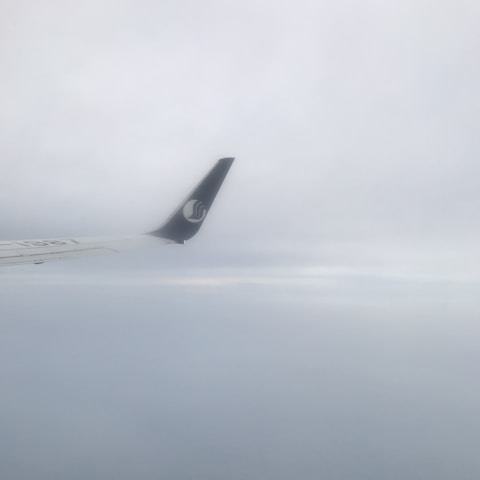 An airplane wingtip is visible against a backdrop of cloudy skies. The sky appears overcast, with varying shades of grey and white.