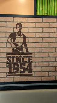 A brick wall features a stencil-style artwork depicting a person wearing an apron beside the text 'Since 1954'. Green bamboo stalks are integrated into the wall's design, adding a touch of nature.