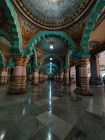 An intricately designed interior features a series of ornate arches and columns with detailed carvings and patterns. The arches are painted in vibrant turquoise and gold hues, with a polished marble floor reflecting the ceiling's ornate details. The lighting gives a soft glow, enhancing the grandeur of the space.