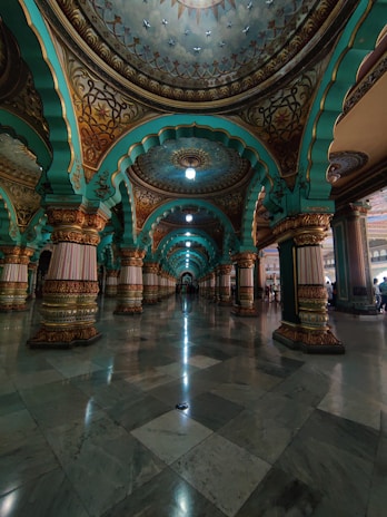 An intricately designed interior features a series of ornate arches and columns with detailed carvings and patterns. The arches are painted in vibrant turquoise and gold hues, with a polished marble floor reflecting the ceiling's ornate details. The lighting gives a soft glow, enhancing the grandeur of the space.