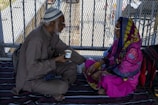 An elderly man and woman sit facing each other on a blanket. The man, dressed in brown attire and wearing a cap, holds a cup, while the woman, in a vibrant pink traditional outfit with a colorful headscarf, listens attentively. They are seated on the ground in an area enclosed by a metal fence, with backpacks nearby.