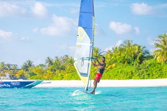 A windsurfer glides across turquoise waters near a lush, tropical island. In the background, a speedboat is visible, and the scene is framed by palm trees and a bright blue sky with fluffy clouds.