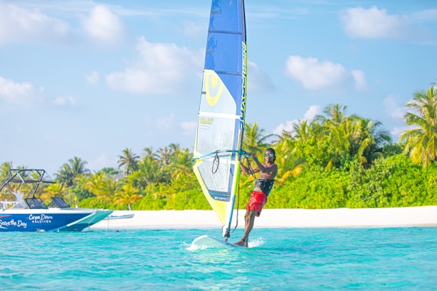 A windsurfer glides across turquoise waters near a lush, tropical island. In the background, a speedboat is visible, and the scene is framed by palm trees and a bright blue sky with fluffy clouds.