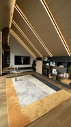 A modern interior space features slanted wooden ceilings with embedded linear lights. A large TV is mounted on a wall with vertical stripes. A contemporary rug with shades of beige, gray, and orange covers the floor. There are open shelving units displaying books and small decor items.