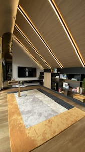 A modern interior space features slanted wooden ceilings with embedded linear lights. A large TV is mounted on a wall with vertical stripes. A contemporary rug with shades of beige, gray, and orange covers the floor. There are open shelving units displaying books and small decor items.
