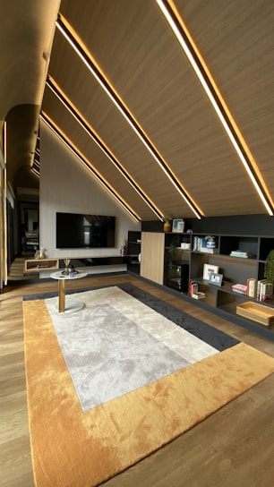 A modern interior space features slanted wooden ceilings with embedded linear lights. A large TV is mounted on a wall with vertical stripes. A contemporary rug with shades of beige, gray, and orange covers the floor. There are open shelving units displaying books and small decor items.