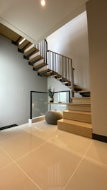 A modern interior featuring an open wooden staircase with metal railings, leading up against a light-colored wall. The floor is polished, light tile, reflecting natural light. Beside the stairs, there is a small, cushioned ottoman and a potted plant on a glass table.