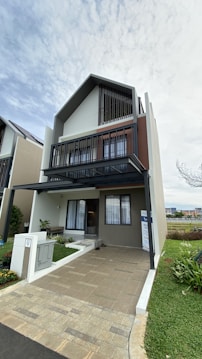 A modern, three-story house features a geometric design with large windows and clean lines. The exterior includes neutral colors such as beige and grey, with dark accents. A small, well-maintained garden is visible at the front, along with a paved driveway. The sky is overcast, adding a soft lighting effect to the scene.