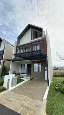 A modern, three-story house features a geometric design with large windows and clean lines. The exterior includes neutral colors such as beige and grey, with dark accents. A small, well-maintained garden is visible at the front, along with a paved driveway. The sky is overcast, adding a soft lighting effect to the scene.
