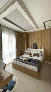 A modern bedroom with a comfortable bed featuring white linens and decorative pillows, surrounded by wooden paneling on the walls. A large window with sheer curtains allows natural light to fill the room. A bedside table holds a lamp, and a tray with cups sits on the bed. The ceiling has a recessed design with spotlights.