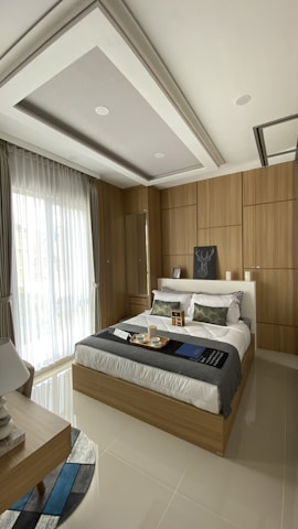 A modern bedroom with a comfortable bed featuring white linens and decorative pillows, surrounded by wooden paneling on the walls. A large window with sheer curtains allows natural light to fill the room. A bedside table holds a lamp, and a tray with cups sits on the bed. The ceiling has a recessed design with spotlights.