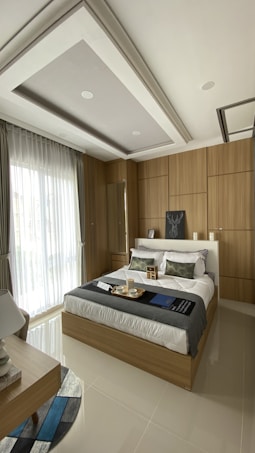 A modern bedroom with a comfortable bed featuring white linens and decorative pillows, surrounded by wooden paneling on the walls. A large window with sheer curtains allows natural light to fill the room. A bedside table holds a lamp, and a tray with cups sits on the bed. The ceiling has a recessed design with spotlights.