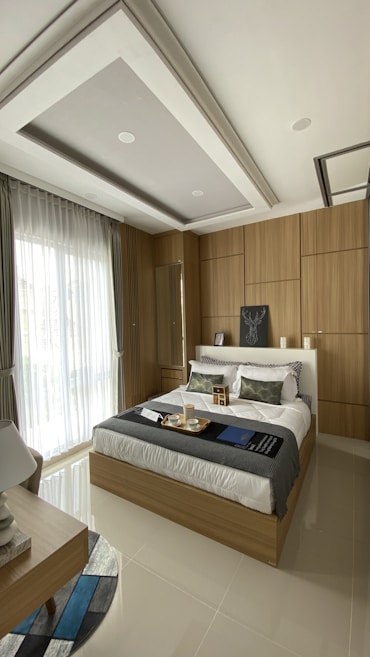 A modern bedroom with a comfortable bed featuring white linens and decorative pillows, surrounded by wooden paneling on the walls. A large window with sheer curtains allows natural light to fill the room. A bedside table holds a lamp, and a tray with cups sits on the bed. The ceiling has a recessed design with spotlights.