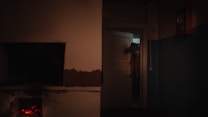 A dimly lit room with a door slightly ajar reveals an ominous figure wearing what appears to be an animal or bird head mask. In the foreground, there is a partially visible open fireplace with glowing embers, casting a warm but eerie light.