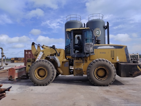 A large yellow construction vehicle, resembling a front loader, is parked on a concrete surface. A person is seated inside the cabin, which has transparent windows. Two large cylindrical tanks are visible in the background, along with a clear blue sky.