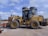 A large yellow construction vehicle, resembling a front loader, is parked on a concrete surface. A person is seated inside the cabin, which has transparent windows. Two large cylindrical tanks are visible in the background, along with a clear blue sky.