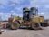 A large yellow construction vehicle, resembling a front loader, is parked on a concrete surface. A person is seated inside the cabin, which has transparent windows. Two large cylindrical tanks are visible in the background, along with a clear blue sky.