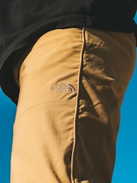 A close-up shot of a person wearing mustard-colored pants with 'The North Face' logo. The pants are in focus against a vibrant blue background, partially showing a black shirt above.