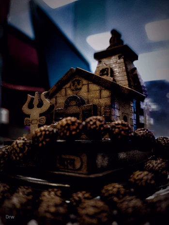 A miniature wooden structure depicting a house or temple with intricate carvings is surrounded by a circle of small brown objects resembling seeds or nuts. The background shows a blurred interior space with subdued lighting, creating a mysterious and moody atmosphere.