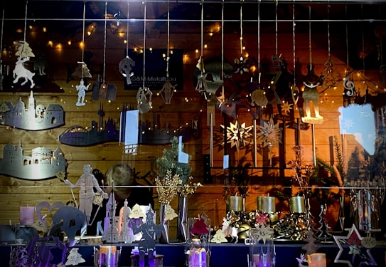 A shop window display featuring various metal ornaments hanging from strings. The ornaments include figures, stars, musical symbols, and architectural shapes. The background has a warm wooden texture, and string lights create a cozy atmosphere. Additional festive decorations, such as small trees and candles, are displayed at the bottom.