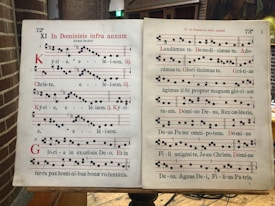 A two-page spread of a Gregorian chant manuscript, featuring black square musical notes on a four-line staff with red lines. The text is in Latin, and the pages are aged and slightly yellowed. The manuscript is open on a wooden lectern in a brick interior space.