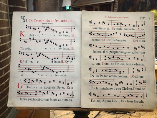 A two-page spread of a Gregorian chant manuscript, featuring black square musical notes on a four-line staff with red lines. The text is in Latin, and the pages are aged and slightly yellowed. The manuscript is open on a wooden lectern in a brick interior space.