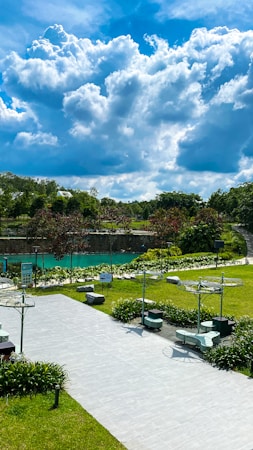 A serene park landscape featuring a well-maintained grassy area with scattered benches and modern lamp posts. In the background, there is a lush row of trees accompanied by a turquoise water body. The sky is dominated by large, fluffy cloud formations against a vibrant blue.