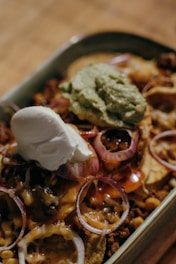 A dish filled with nachos topped with melted cheese, red onion slices, guacamole, and sour cream. The ingredients are layered to create a visually appealing and appetizing meal in a rectangular dish.