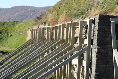 An outdoor scene with a stone retaining wall supported by wooden beams, set against a green grassy hillside. The background features a range of hills or mountains with autumn foliage.