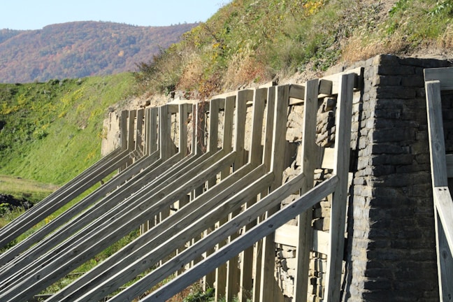 An outdoor scene with a stone retaining wall supported by wooden beams, set against a green grassy hillside. The background features a range of hills or mountains with autumn foliage.