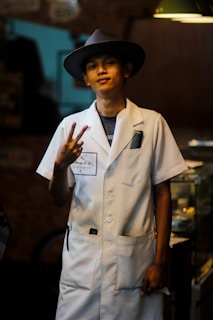 A person wearing a white coat and a wide-brimmed hat, standing indoors with a relaxed expression. They are making a peace sign gesture with their fingers. The background is dimly lit with some furniture partially visible.