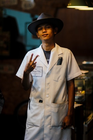 A person wearing a white coat and a wide-brimmed hat, standing indoors with a relaxed expression. They are making a peace sign gesture with their fingers. The background is dimly lit with some furniture partially visible.