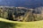 A tranquil hillside landscape with a mix of dense green and autumnal trees stretching across the scene. A small cabin with a sloping roof is nestled within the trees, surrounded by a lush grassy meadow. The distant mountains and a soft light give the area a peaceful, secluded feel.